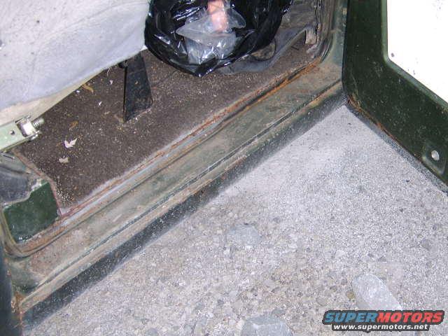 Interior shot of a vehicle's door sill, showing rust and dirt. A black plastic bag is on the floor mat