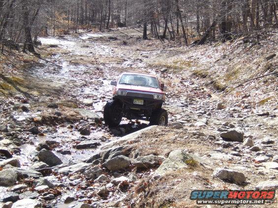 Red SUV with large tires navigating a rocky, wet trail in a wooded area
