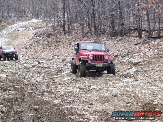 Red Jeep Wrangler driving on a rocky, wooded trail. Another red SUV is visible further up the path