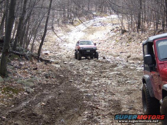 Red Jeep and another off-road SUV on a muddy, rocky trail in a wooded area