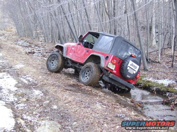 Red Jeep Wrangler with a soft top driving off-road on a muddy path with trees and snow patches