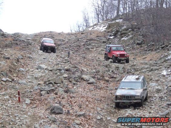 Three SUVs are driving up a steep, rocky hill. One is red, one is camouflaged, and one is dark red