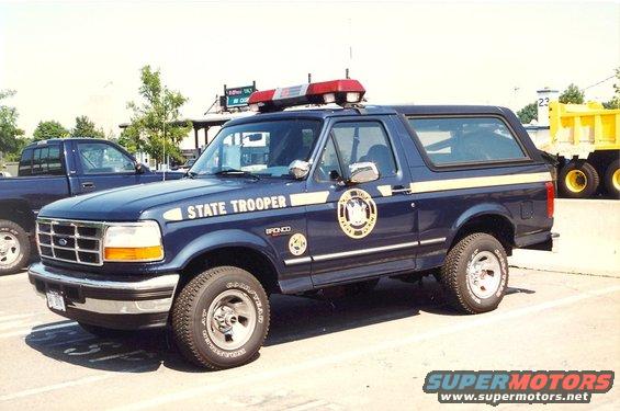 Dark blue Ford Bronco with "STATE TROOPER" on the side, light bar on top, parked next to a dark blue pickup truck