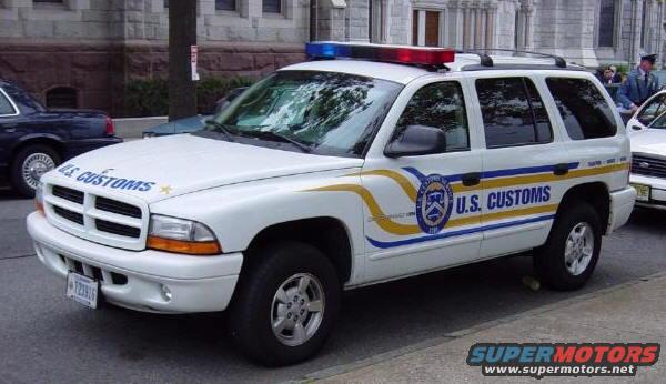 White U.S. Customs SUV with light bar on roof parked next to a dark sedan and another white car