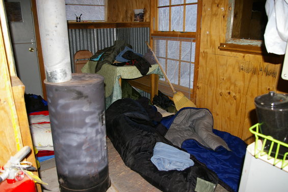 Interior of a cabin with a wood stove, sleeping bags, and a table with clothes