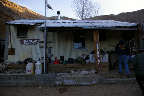 Briggs Mine Cabin with corrugated metal siding and a sign reading 'BRIGGS MINE CABIN Adopt-a-Cabin'. Two people stand near the entrance