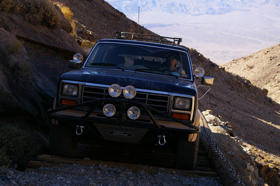 Dark blue Ford Bronco driving on a narrow, rocky mountain path with a driver visible inside. It has a roof rack and extra lights on the front