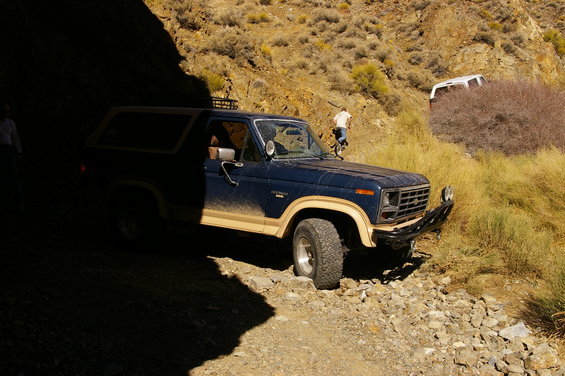 Dark blue Ford Bronco with tan trim, mud-splattered, parked on a rocky path