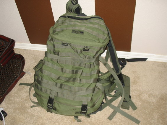 Green tactical backpack with MOLLE webbing and black buckles, shown from the front on a carpeted floor