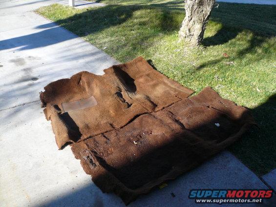 Brown car carpet laid out on concrete and grass, showing wear and tear