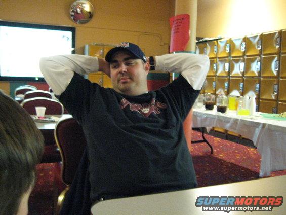 Man with hands behind head, wearing a dark t-shirt and baseball cap, sits at a table