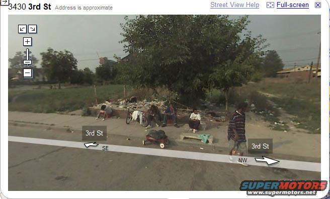 People sitting and standing on a sidewalk next to a street, with trash and trees in the background
