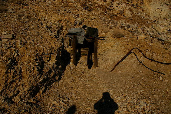 Mine entrance with wooden supports and rusty metal, set in a rocky, arid landscape. Shadow of a person is visible