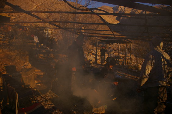 People gathered under a wooden structure, with smoke rising from a fire in the foreground. Mountains are visible in the background