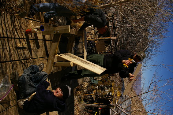 three people working on a wooden structure outdoors, one is using a drill