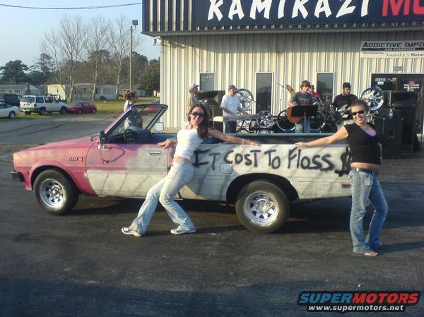 Two women pose with a pink and white pickup truck that has graffiti on its side. A band plays in the background