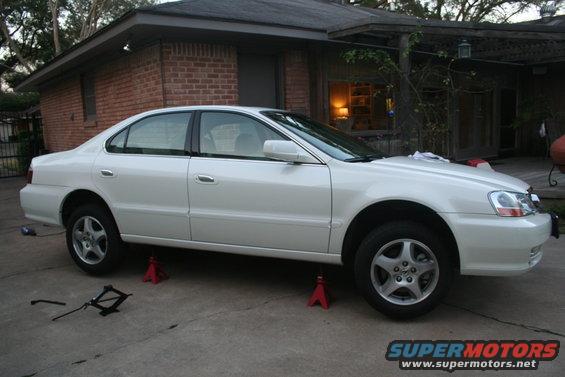 White 2003 Acura TL on jack stands in a driveway with a jack and lug wrench on the ground