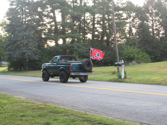 Green Ford Bronco driving on a two-lane road, spare tire mounted on the rear, Confederate flag flying from a pole