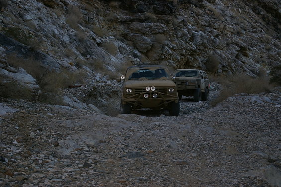 Two modified SUVs driving on a rough, rocky mountain trail. The lead vehicle has a custom bumper and extra lights
