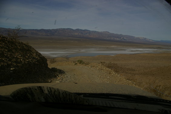 View from inside a vehicle looking out at a vast desert landscape with mountains in the distance and a dry lake bed