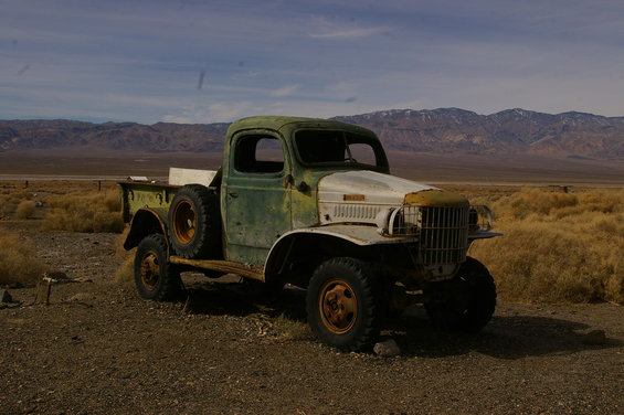 Green and white weathered truck with a spare tire on the side, parked on a dirt road with dry grass and mountains in the background