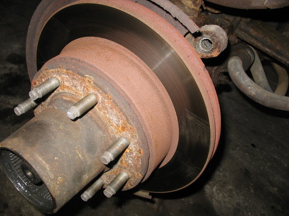 Close-up of a heavily rusted wheel hub with five lug studs and a brake rotor. Suspension components are visible in the background