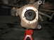 1995 Ford C-350 (Centurion Conversions) - Close-up of a rusty front axle spindle, supported by a red jack stand, with the vehicle's undercarriage visible above.