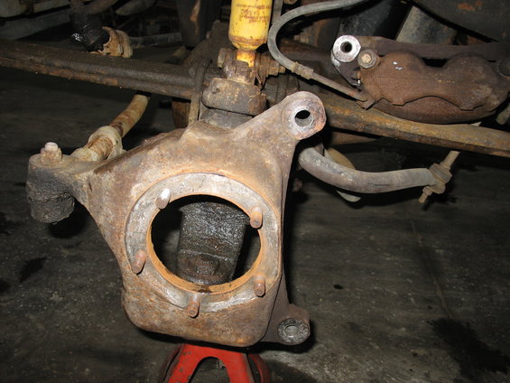 Rusty front axle spindle assembly on red jack stand, showing brake caliper bracket and tie rod end