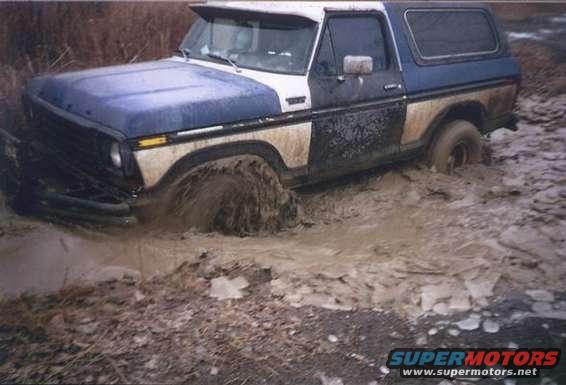 Blue and white Ford Bronco stuck in mud, front wheel deep, side view