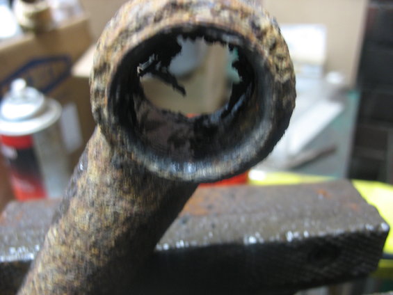 Rusted metal pipe section held in vise, showing severe internal corrosion and damage