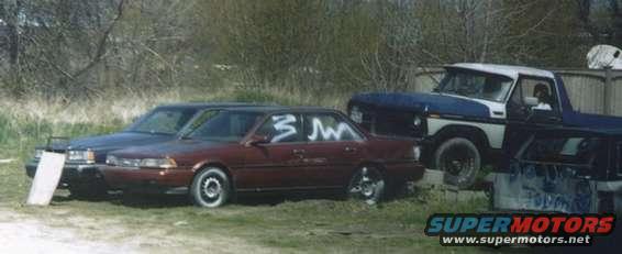 Several cars and a pickup truck parked in a grassy area with trees in the background. White markings on the side of a red sedan
