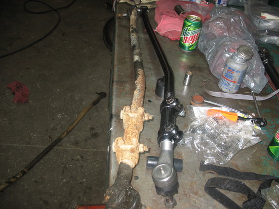 Old rusty steering linkage next to a new black steering linkage on a workbench