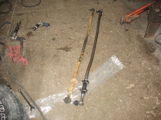 Two tie rods, one rusty and one new, lie on a concrete floor next to tools and a tire