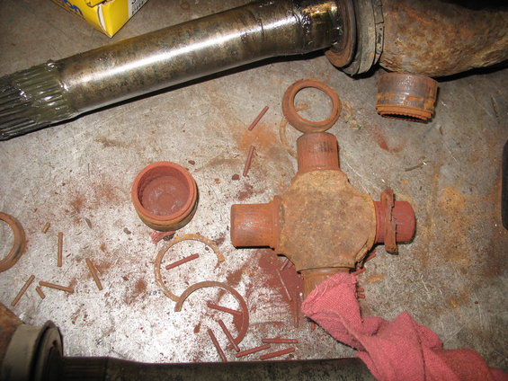 Driveshaft universal joint and axle components disassembled on a metal surface. Red dust covers the parts