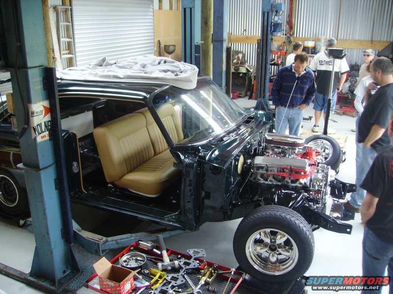 Car in repair shop, interior visible, engine bay empty, some people standing around