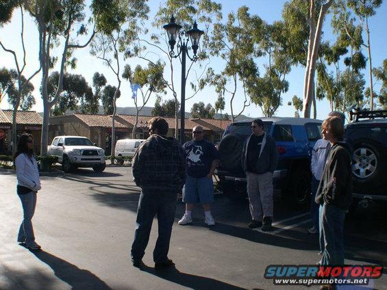 Several people stand around a few SUVs in a parking lot on a sunny day
