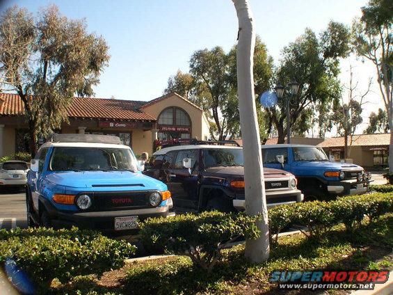 Three Toyota FJ Cruisers parked, one blue, one dark purple, one light blue. Front view