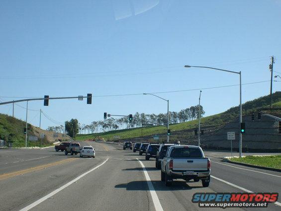 Traffic intersection with several vehicles in lanes, including pickup trucks and SUVs. Clear blue sky above a grassy hill