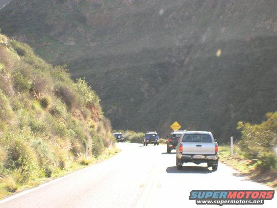 Several trucks and SUVs driving on a road next to a hill. A yellow diamond sign is visible
