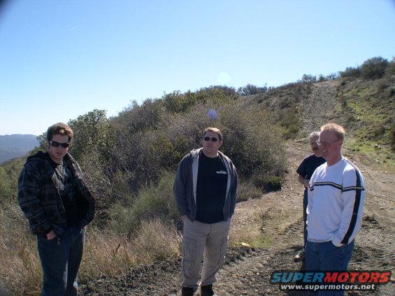 Four men stand on a dirt road with hills and brush in the background
