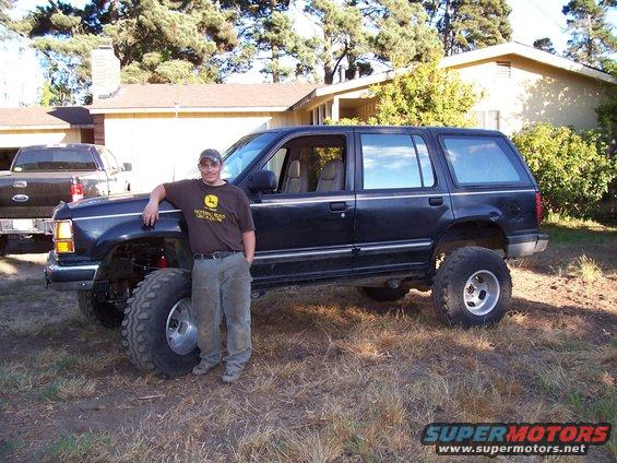Man leaning on a lifted dark blue Ford Explorer with large tires. Another truck is partially visible to the left