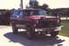 1984 Ford Bronco - This red SUV features aftermarket lights mounted on the hood, seen from a 3/4 front angle.