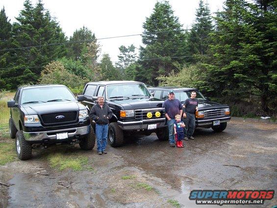 Four black Ford trucks and SUVs parked on a wet, gravelly surface with trees in the background