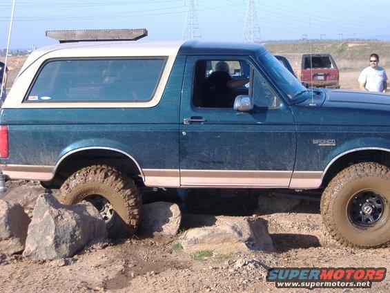 Green Ford Bronco with white top navigating rocks on a dirt trail. Another SUV and a person are visible in the background