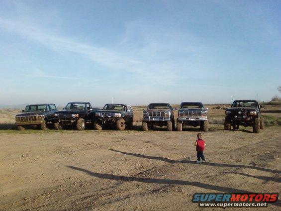Several lifted Ford trucks and SUVs parked in a dirt lot under a clear sky