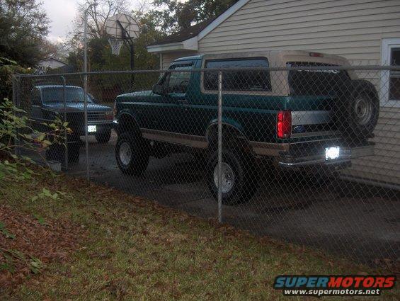 Green Ford Bronco with tan top and spare tire on rear, parked behind a chain-link fence