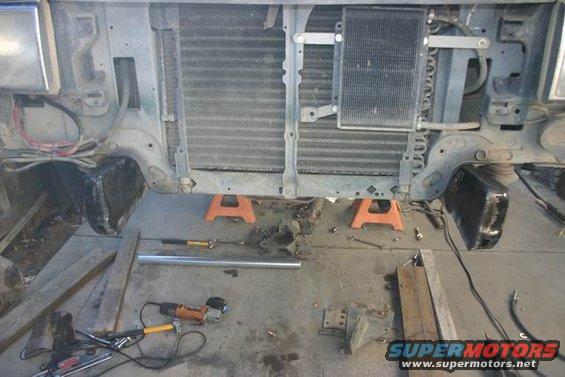 Front end of a vehicle disassembled, with radiator and AC condenser exposed. Tools and jack stands are on the ground