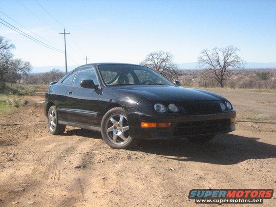 Black coupe parked on dirt road, trees and hills in background