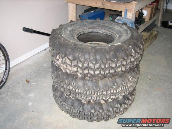 stack of three large off-road tires, one on top of two, with a bicycle wheel and handle visible to the left