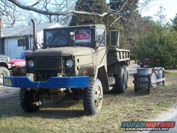 Military truck with blue bumper and 'For Sale' sign in window. Another vehicle is partially visible to the left